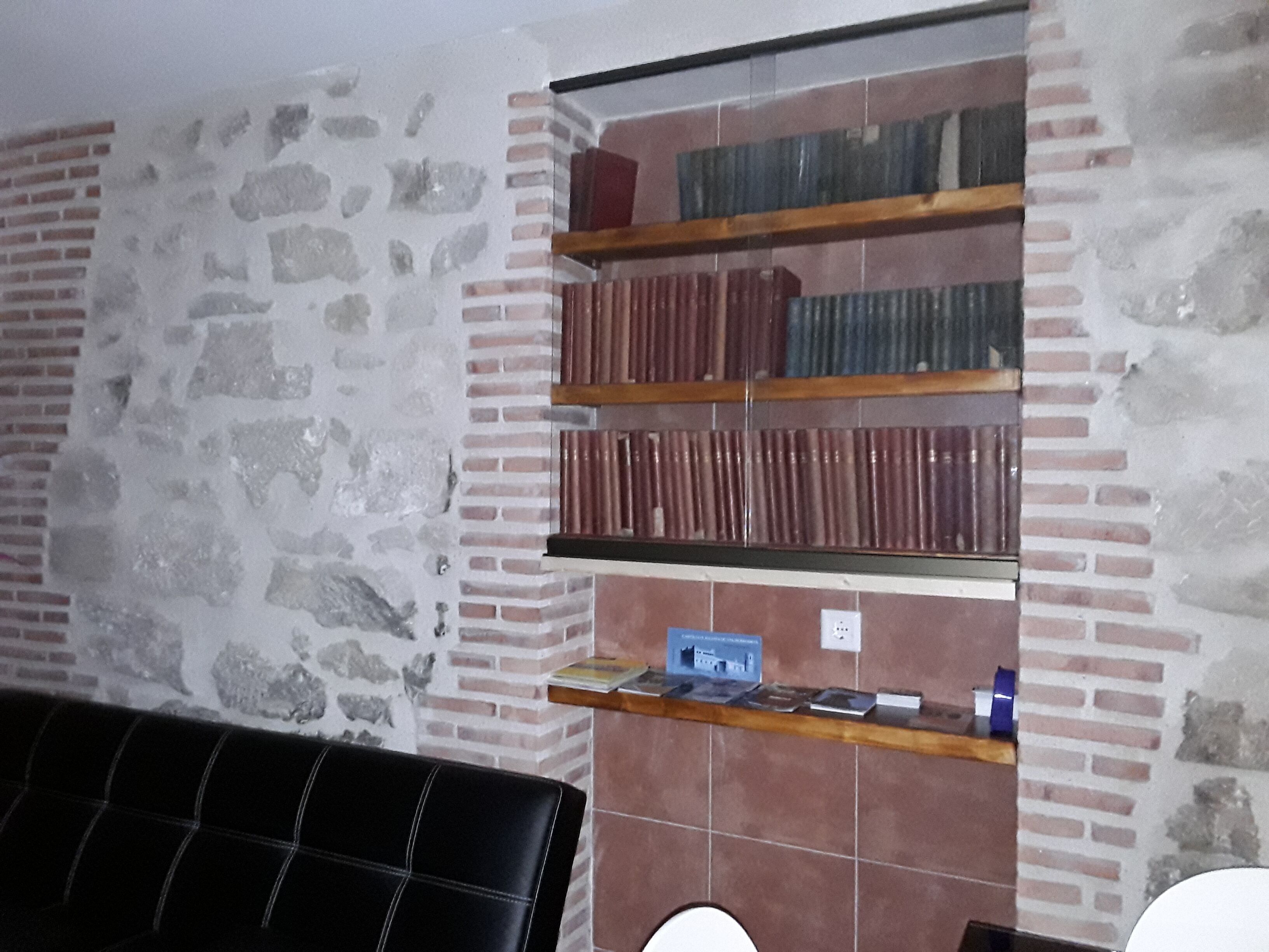 Una habitación con pared de piedra, pared de ladrillo y suelo de baldosas. Hay un sofá de cuero negro y una estantería de madera con libros.