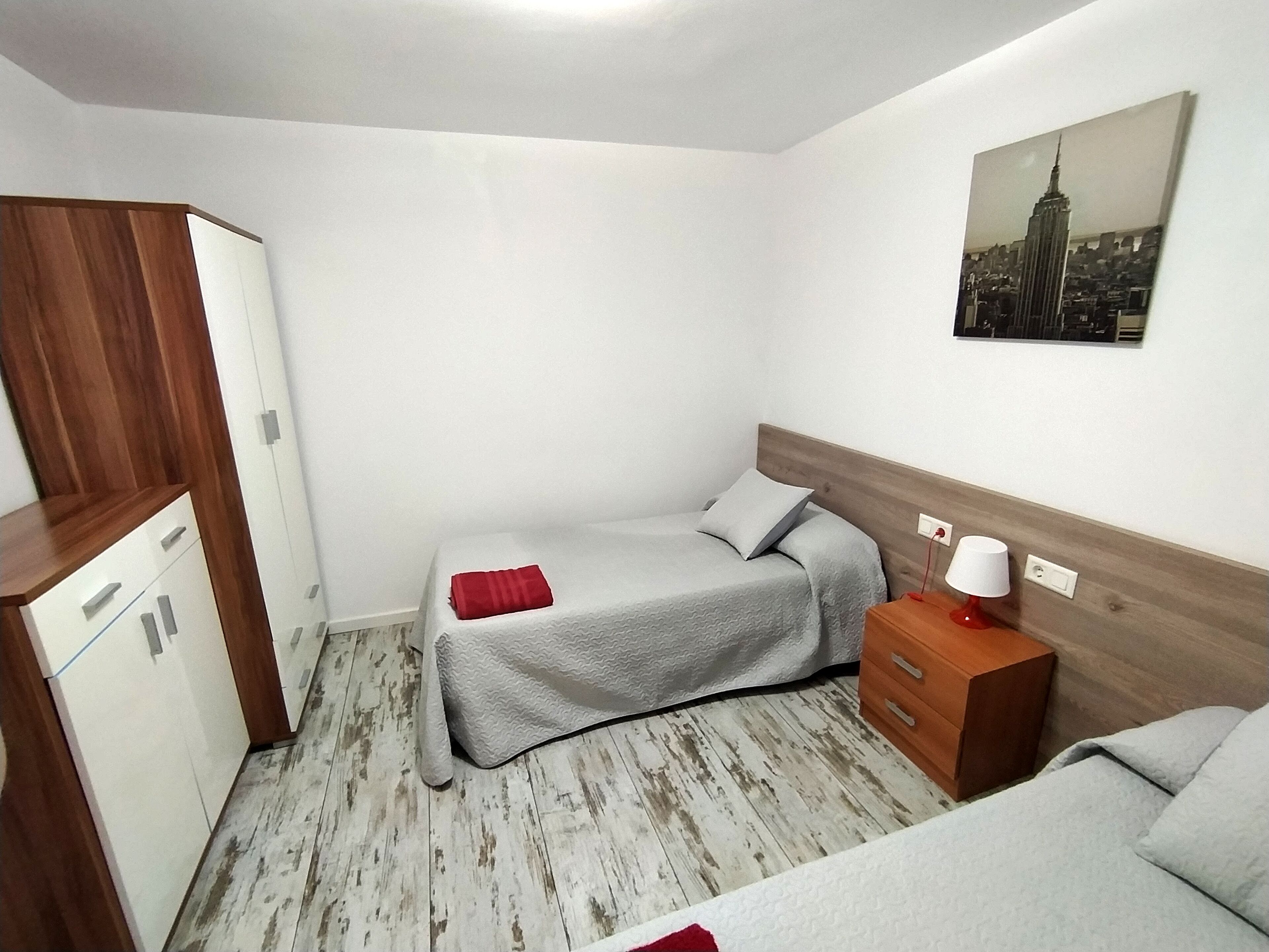 Une chambre d’hôtel de taille modeste, comprenant un lit, une commode en bois, une table de chevet et un tableau représentant un paysage urbain accroché au mur.