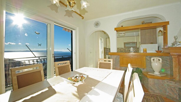A dining area with a table set for two, a view of the sea through large windows, and a balcony with a railing. - Apartment Ana Put Makra (Makarska)