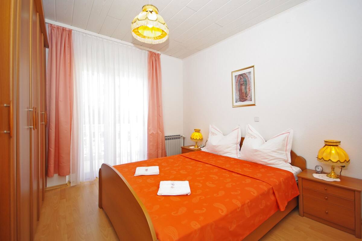 A bedroom with a bed, two bedside tables, a nightstand, a lamp, a radiator, and a window with curtains.