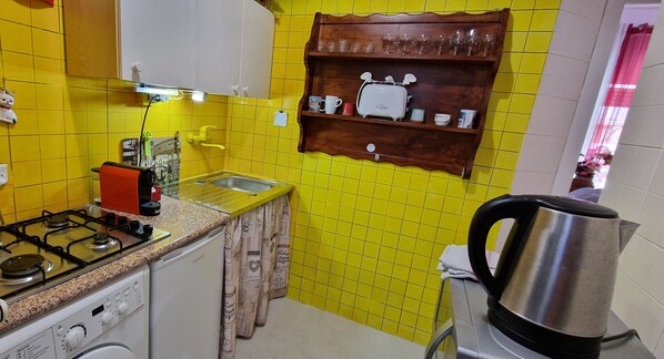 Una cocina con baldosas amarillas, estufa a gas, pileta, estante de madera con utensilios de cocina y tetera.