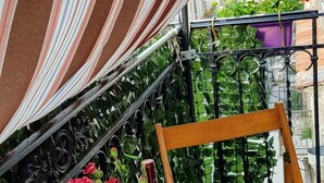 Een houten stoel met een geweven tafeltje op een balkon, met gestreepte stoffen gordijnen, potplanten en uitzicht op gebouwen.