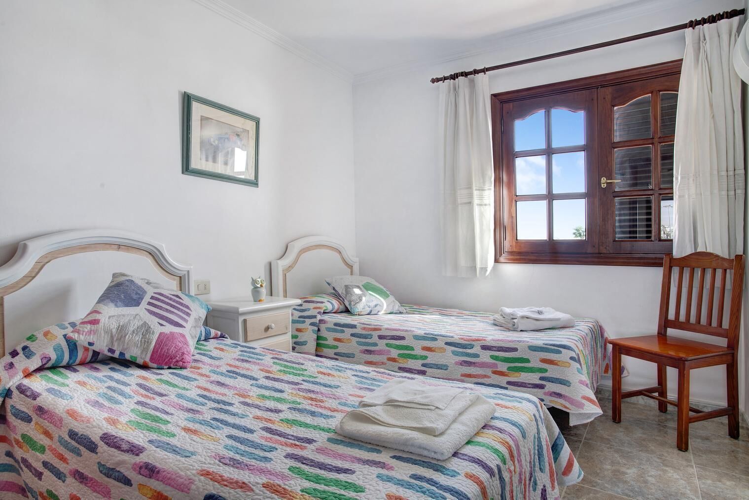 A bedroom with two beds, a wooden chair, a nightstand, a window with curtains, and a framed picture on the wall.