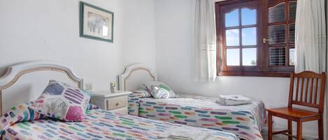 A bedroom with two beds, a wooden chair, a nightstand, a window with curtains, and a framed picture on the wall.