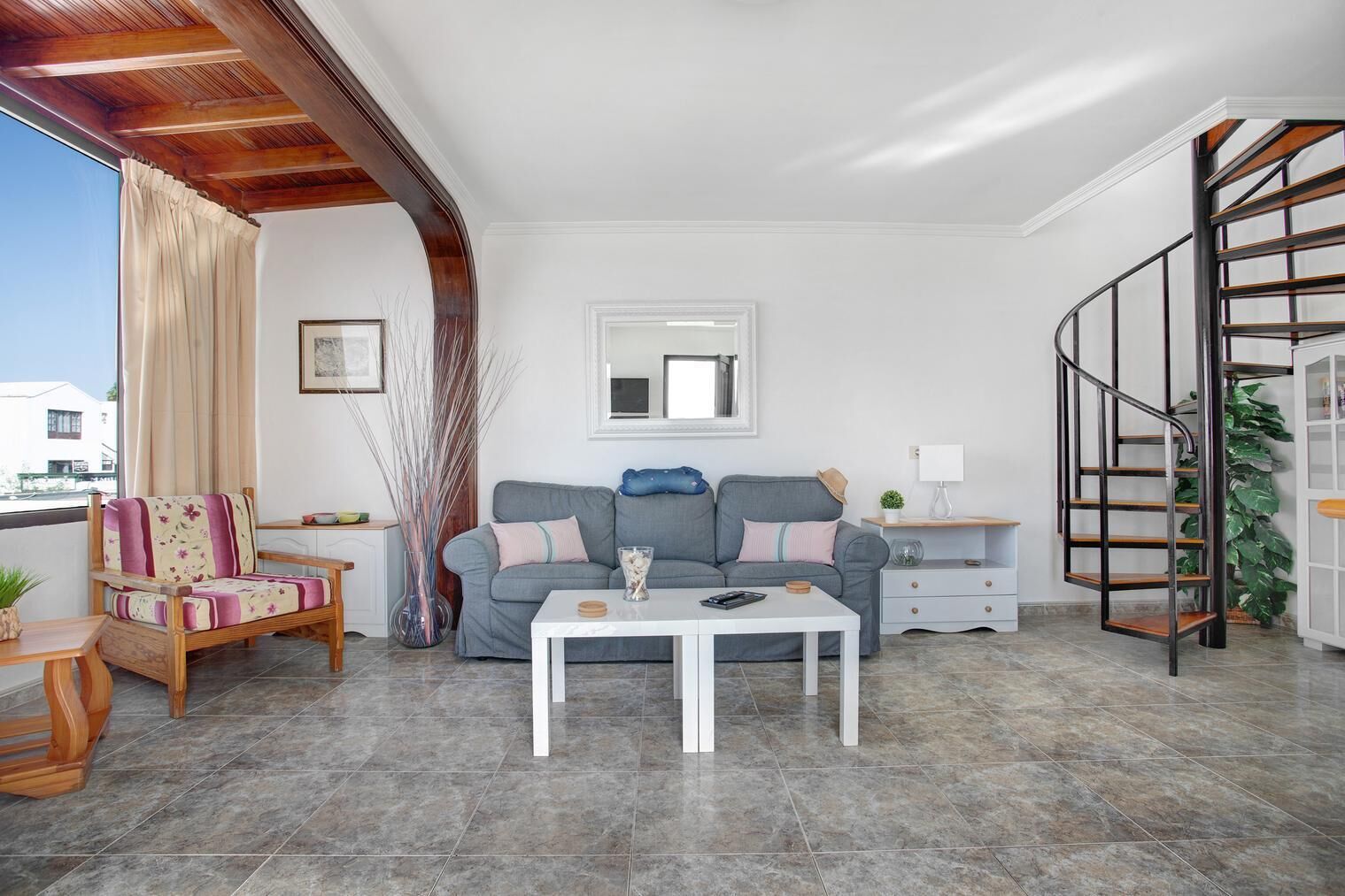 A living room with a grey sofa, a white coffee table, a wooden chair, a small table with a plant, and a staircase.