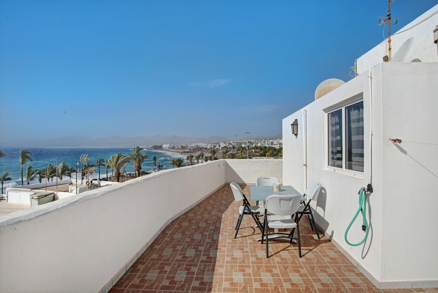A balcony with a table and chairs, a hose, and a view of the ocean and cityscape.