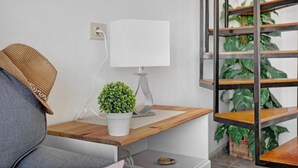 A modern living room with a grey sofa, a wooden side table, a white lamp, a small potted plant, and a bookshelf with green plants. - Apartment the Quintero (Tías)