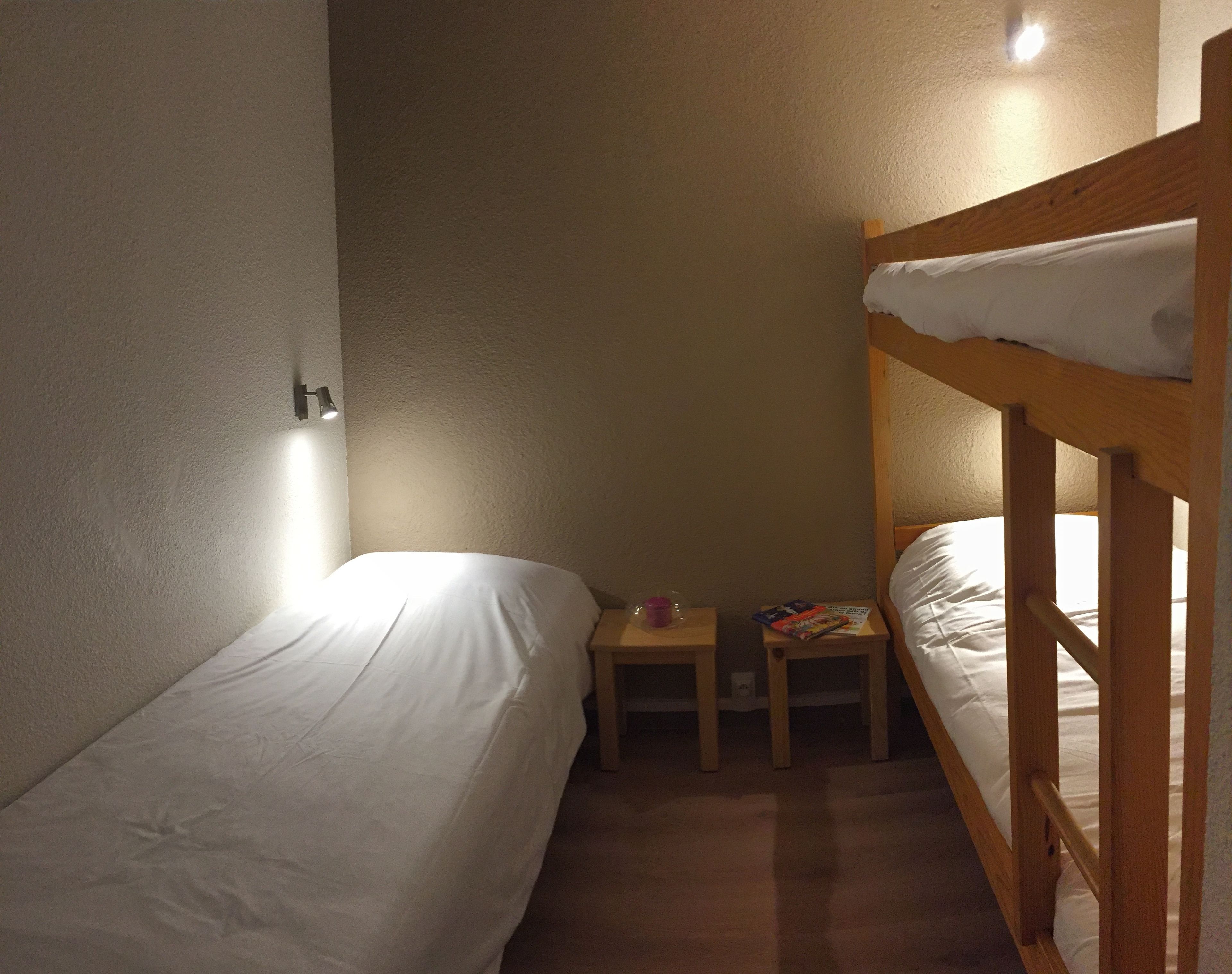 A small, dimly lit room with two bunk beds, a nightstand with a lamp, and a small table with books.