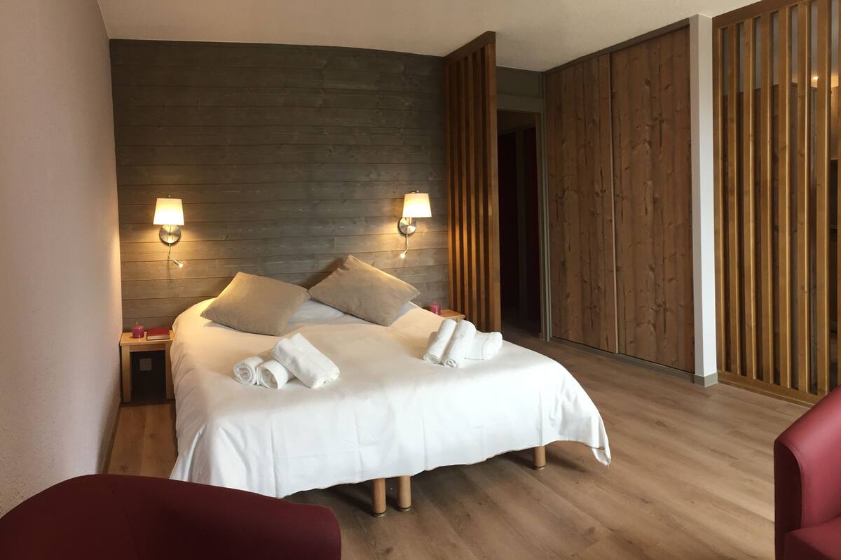A modern hotel room with a wooden headboard, two bedside lamps, a neatly made bed with white linens, and a red chair.