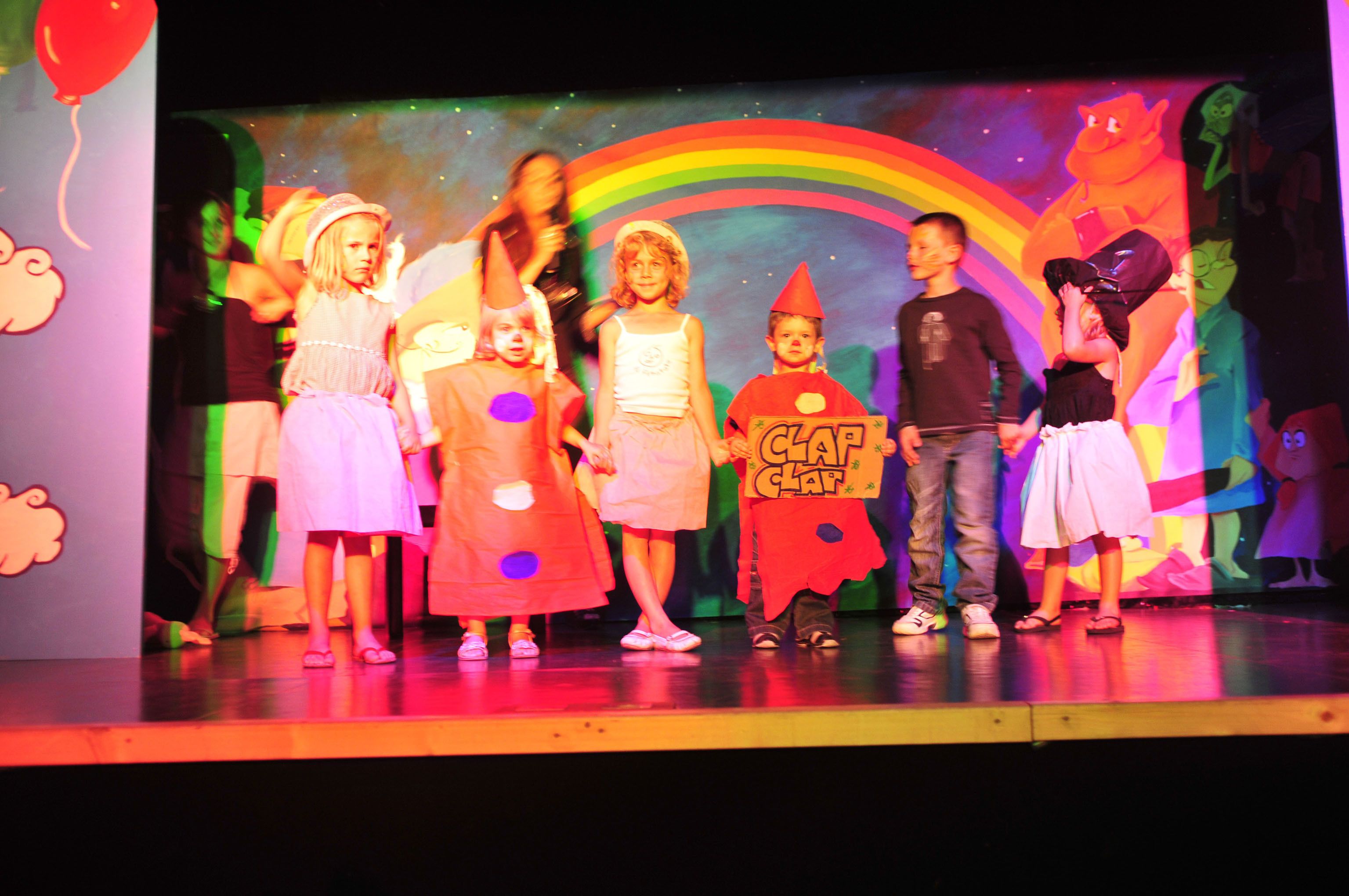 A group of children on a stage with a colorful backdrop featuring a rainbow and clouds.
