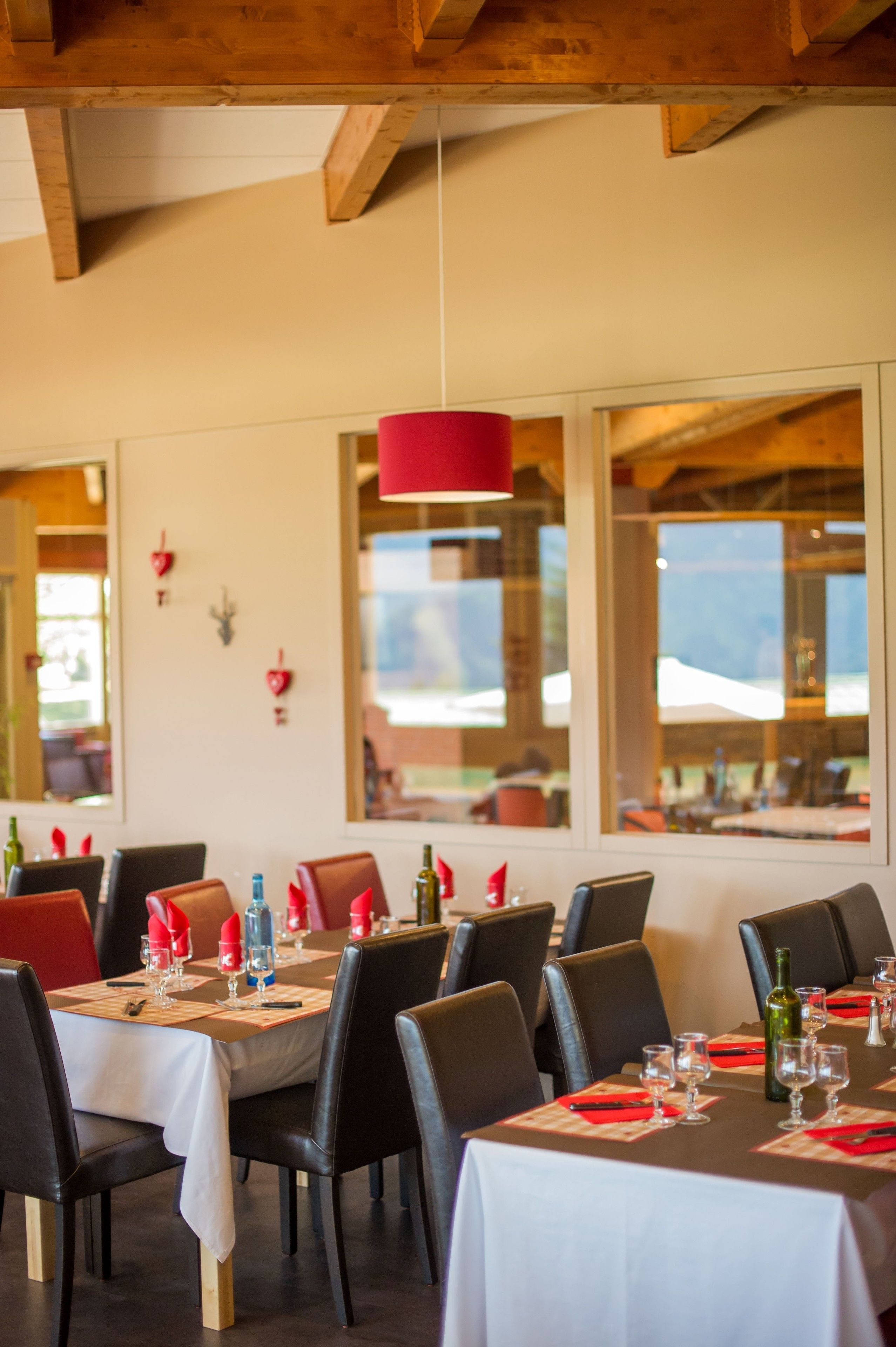 A dining area with tables set for two, featuring a red pendant light, wooden beams, and large windows offering a view of the outdoors.
