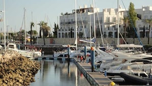 Uma marina com barcos atracados, um cais de madeira e edifícios ao fundo.