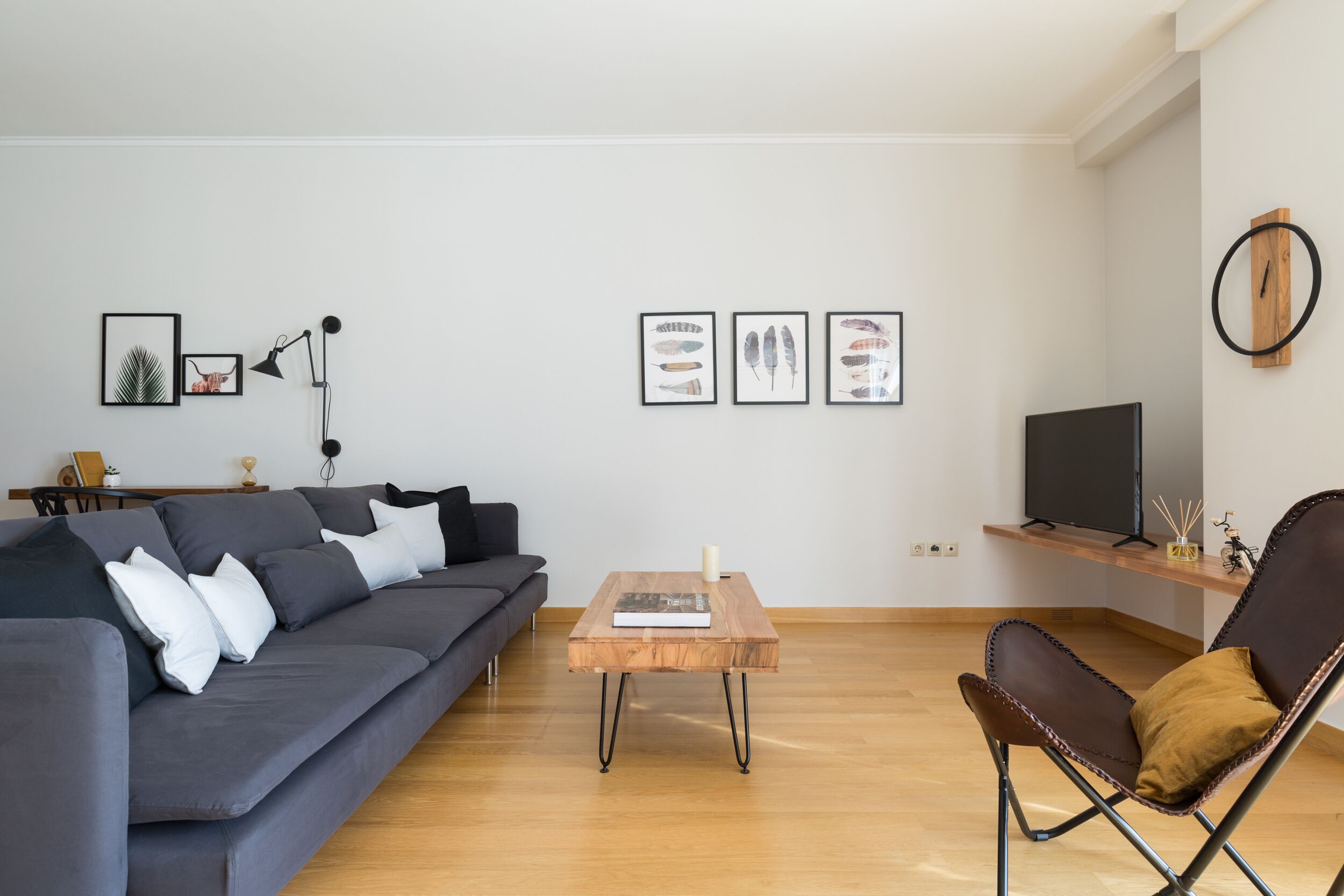 A modern living room with a grey sofa, a wooden coffee table, a leather armchair, and a flat-screen TV on a wooden shelf.