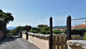 Un carrer estret amb una porta, un senyal amb el número 15, i una vista del mar i les colines al fons.