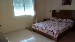 A bedroom with a bed, a nightstand, a window with blinds, and a tiled floor. - Ocean Atlantic Residence 1512 (Asilah)