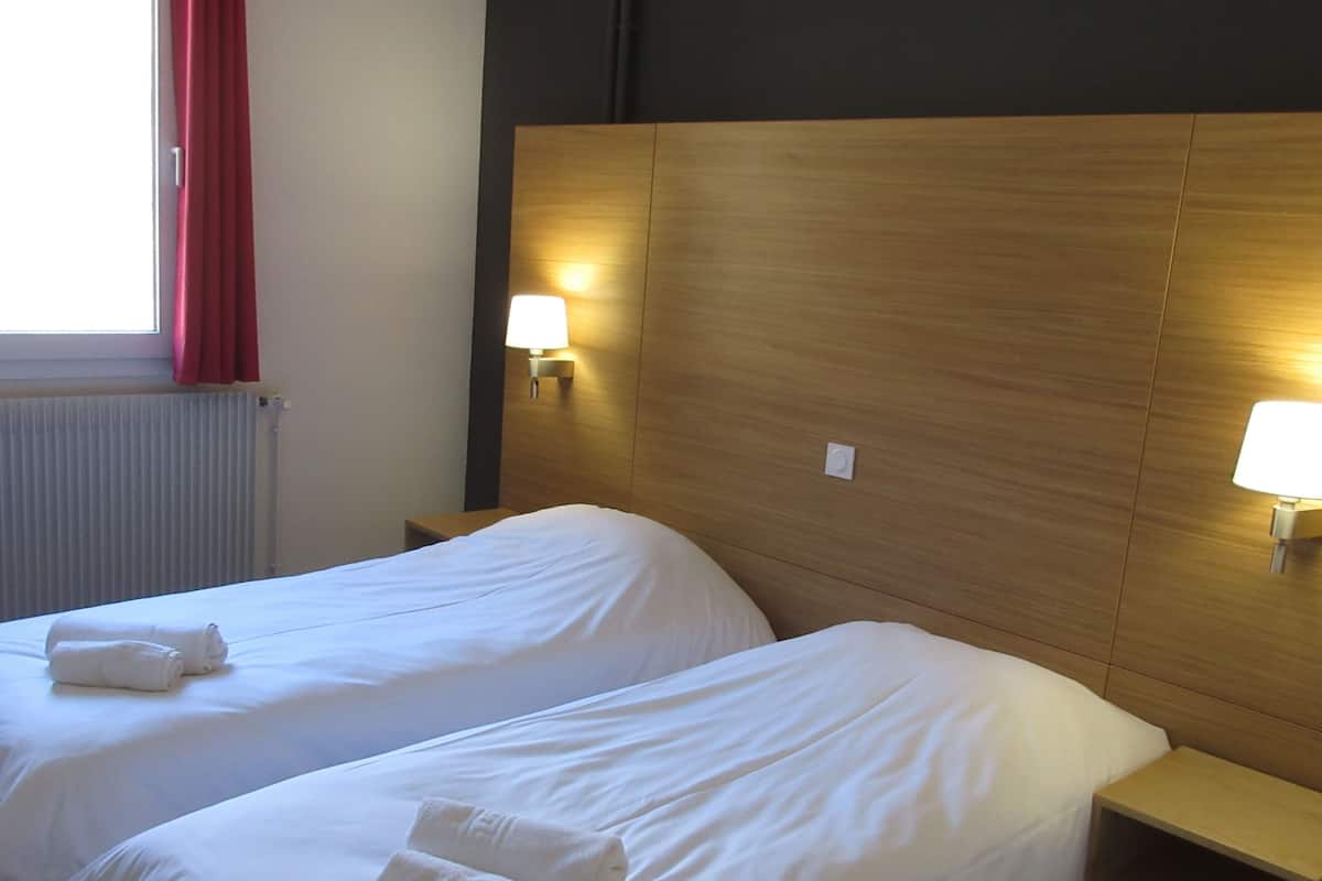 A hotel room with two beds, each with a white duvet and rolled towels. There are bedside lamps on wooden nightstands.
