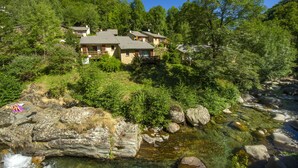 A mountain lodge with multiple buildings surrounded by dense forest and a clear stream. - Village de vacances de Marc - Chambres (Auzat)