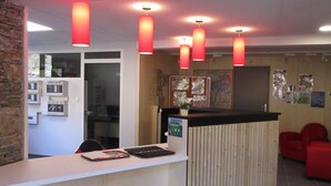 A reception desk with a wooden panel design, a computer, and a plant. There are red pendant lights above the desk. - Village de vacances de Marc - Chambres (Auzat)