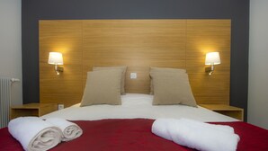 A neatly made bed with a red blanket, two beige pillows, and two white towels folded on the bed. There are two bedside tables with lamps on either side of the bed. - Village de vacances de Marc - Chambres (Auzat)