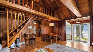 L’intérieur d’une cabane en rondins, avec un escalier en bois, un coin repas comprenant une table et des chaises, une cheminée et une vue à travers une porte vitrée.