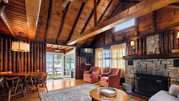 L’intérieur d’un chalet rustique est agrémenté d’une cheminée en pierre, de poutres apparentes au plafond, d’un coin repas avec une table et des chaises, et d’un espace salon confortable avec des canapés et des fauteuils.