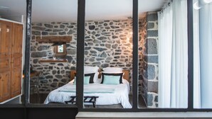A bedroom with a stone wall, wooden bed frame, and a window with curtains. - Aux Caprices d'Aubrac (Laguiole)
