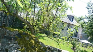 A stone building with a sloped roof, surrounded by lush greenery and a stone wall. - Aux Caprices d'Aubrac (Laguiole)