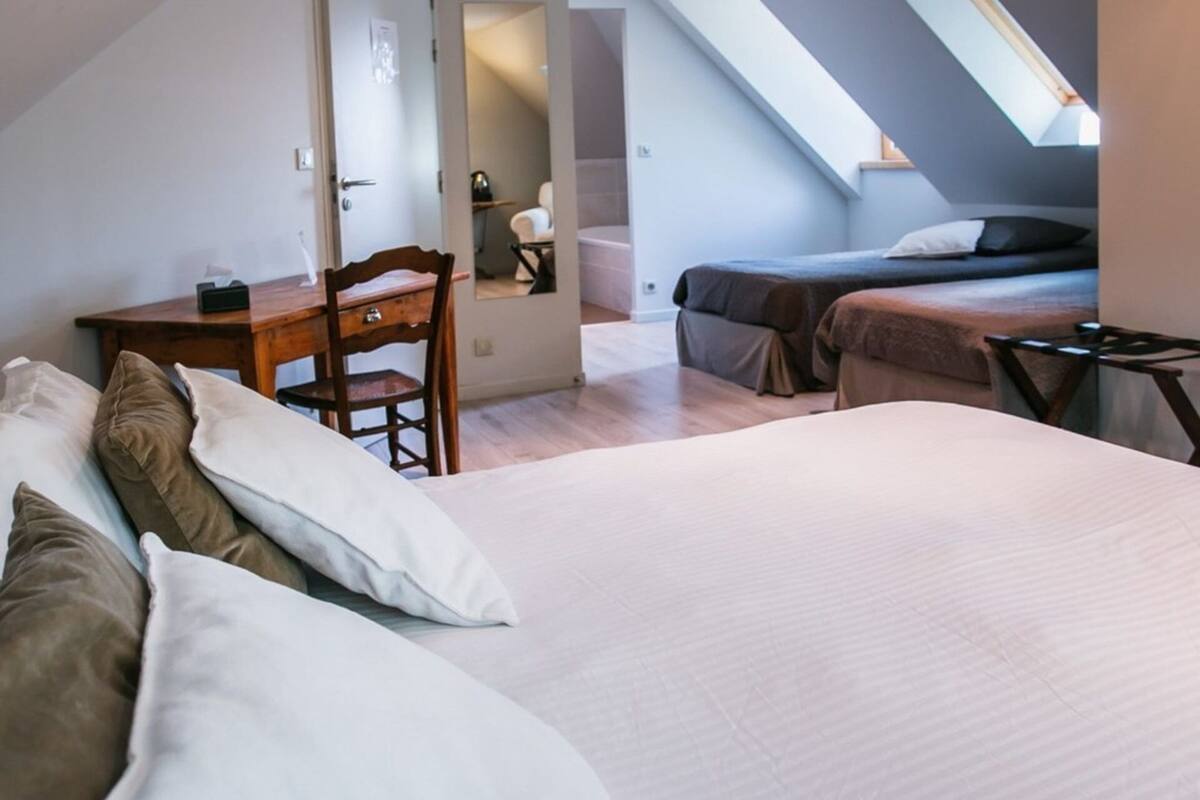 Une chambre avec un grand lit, un bureau et une chaise. On aperçoit un deuxième lit au fond de la pièce et une lucarne au-dessus.