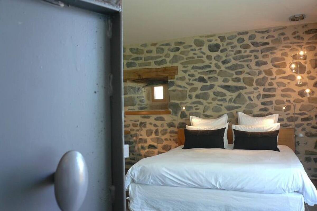 Une chambre avec un mur en pierre, un lit avec une literie blanche et une petite fenêtre.