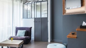 A modern living room with a grey door, a purple armchair, a small table with a vase of flowers, a light blue ottoman, and a mounted television. - Aux Caprices d'Aubrac (Laguiole)