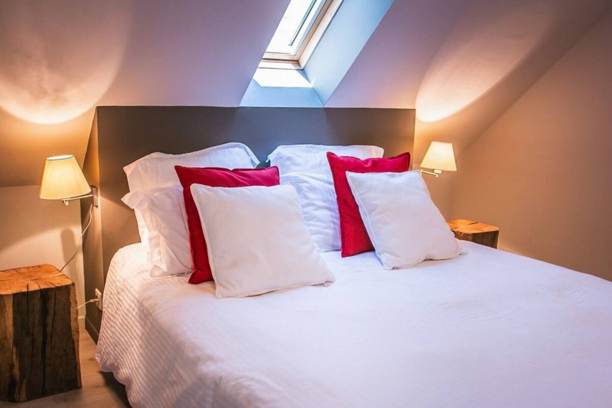 Un lit bien fait, avec des oreillers blancs et rouges, deux lampes de chevet et une lucarne au-dessus.