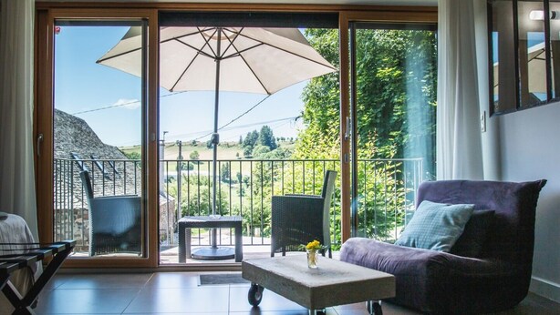 A modern living room with a sliding glass door leading to a balcony, a large umbrella, a purple sofa, and a small table with a vase. - Aux Caprices d'Aubrac (Laguiole)