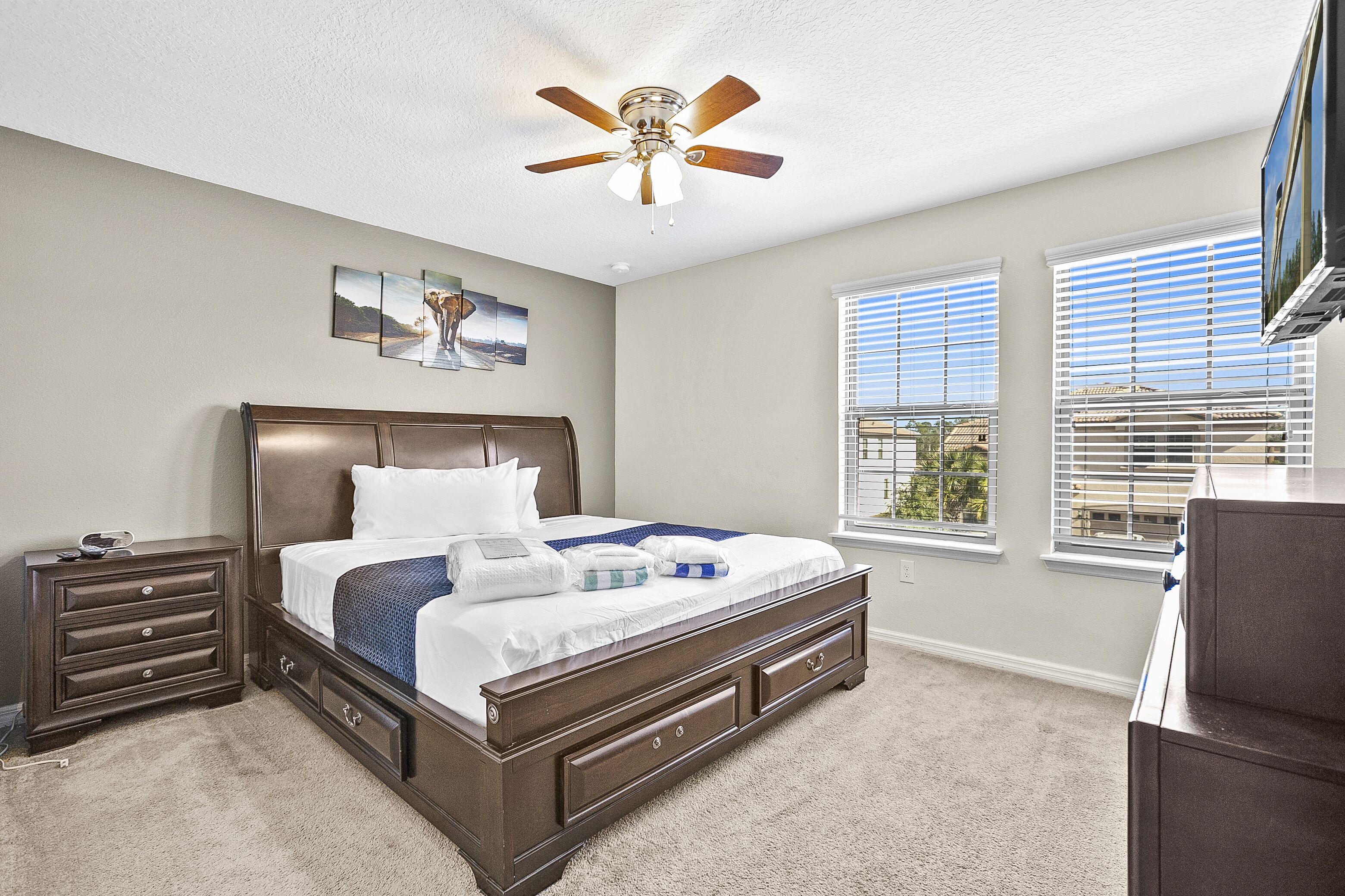 A bedroom with a large bed, a nightstand, a dresser, a ceiling fan, and two windows with blinds.