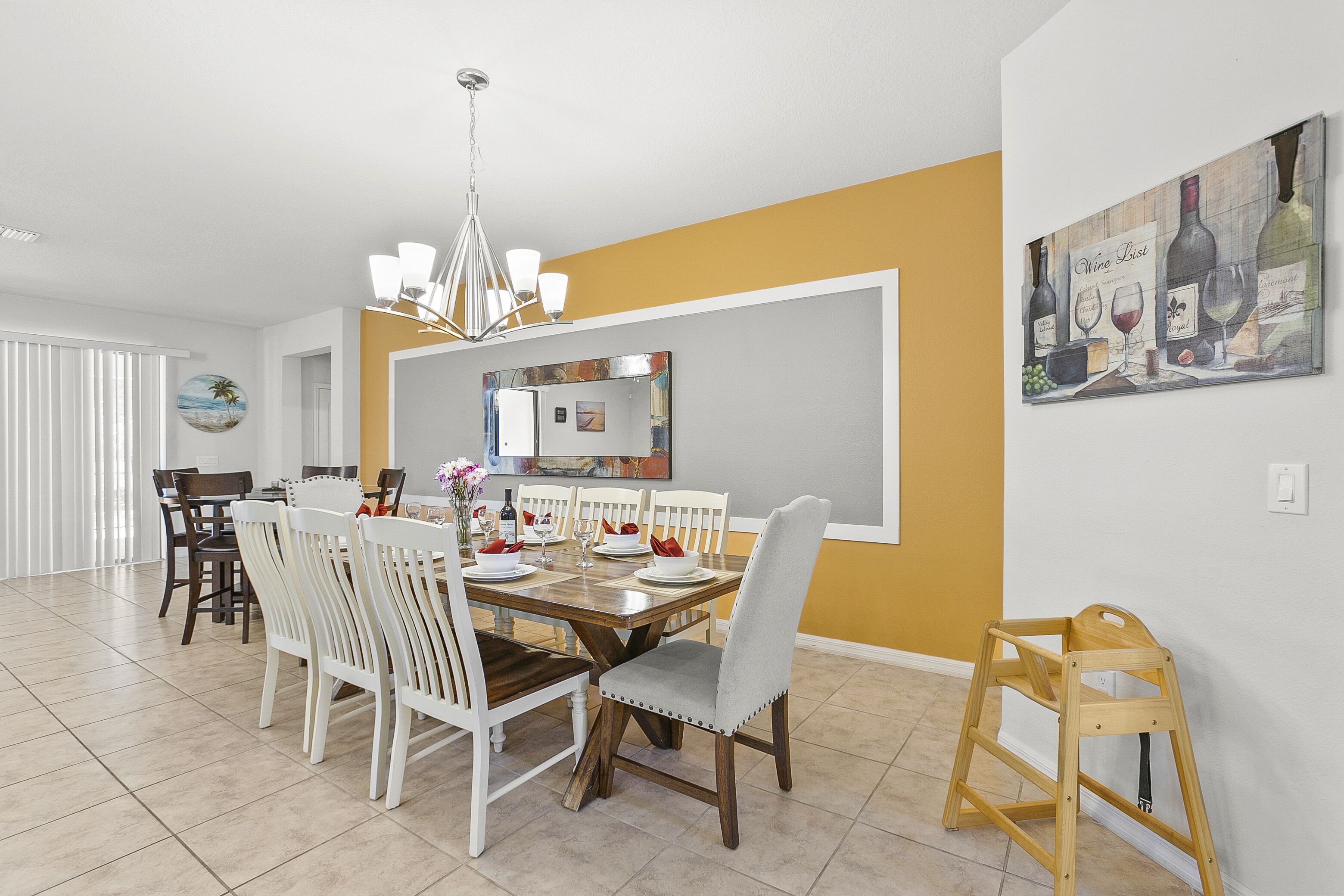 A dining area with a wooden table and white chairs, a chandelier, and a painting on the wall.