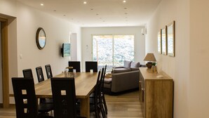 Un comedor amplio con una mesa y sillas de madera, un sofá y vistas al exterior a través de amplias ventanas.