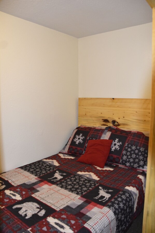 Une petite chambre chaleureuse, avec une tête de lit en bois, une couvre-lit matelassée aux motifs hivernaux et un seul coussin rouge.