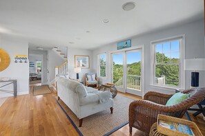 Un salón espacioso con un sofá de tonos claros, un sillón de mimbre y vistas al exterior a través de amplias ventanas.