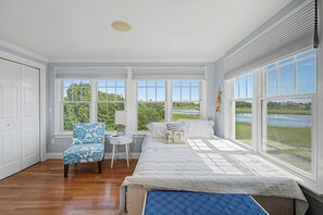 Un dormitorio con cama, una silla, un escritorio y amplios ventanales con vistas a un lago y follaje.