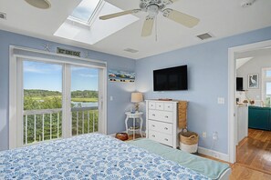 A bedroom with a bed, a ceiling fan, a painting, a television, and a view of the outdoors.