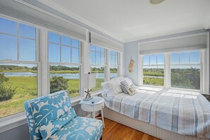 A bedroom with large windows, a bed with a striped bedspread, a blue patterned armchair, and a small bedside table with a lamp.