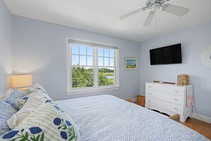 A bedroom with a bed, a dresser, a television, and a window with a view of greenery.