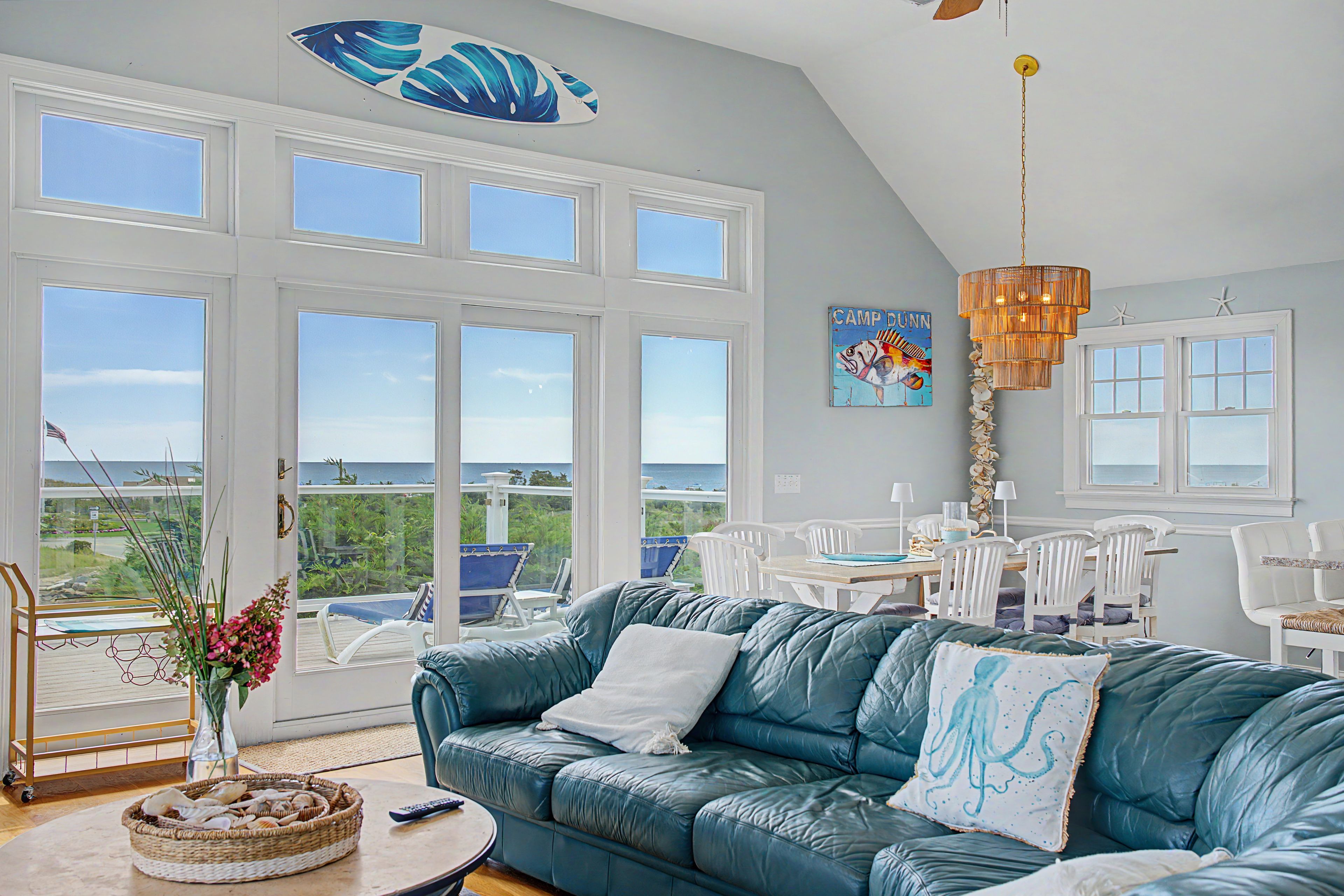 A room with a large window overlooking a scenic view, a teal sofa, a dining area with white chairs, and a decorative ceiling light.