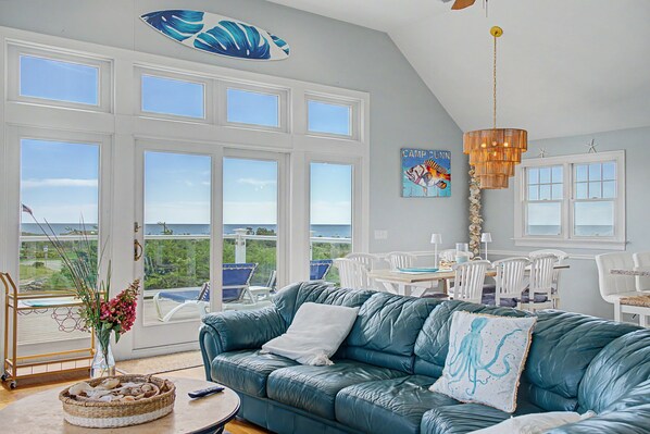 A room with a large window overlooking a scenic view, a teal sofa, a dining area with white chairs, and a decorative ceiling light.