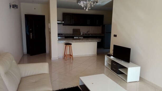 A modern living room with a sofa, a coffee table, a TV on a stand, and a kitchen in the background.