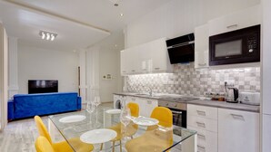 A modern kitchen with white cabinets, a microwave, and a coffee maker. There is a dining area with a glass table and yellow chairs. - Stylish 2 Bedroom Apartment in an Amazing Location (Sliema)