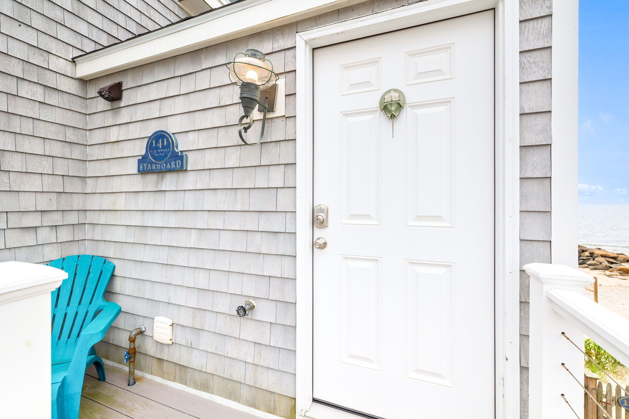 A white door with a green doorknob, a blue street sign reading "141 Starboard," and a blue Adirondack chair on the porch.