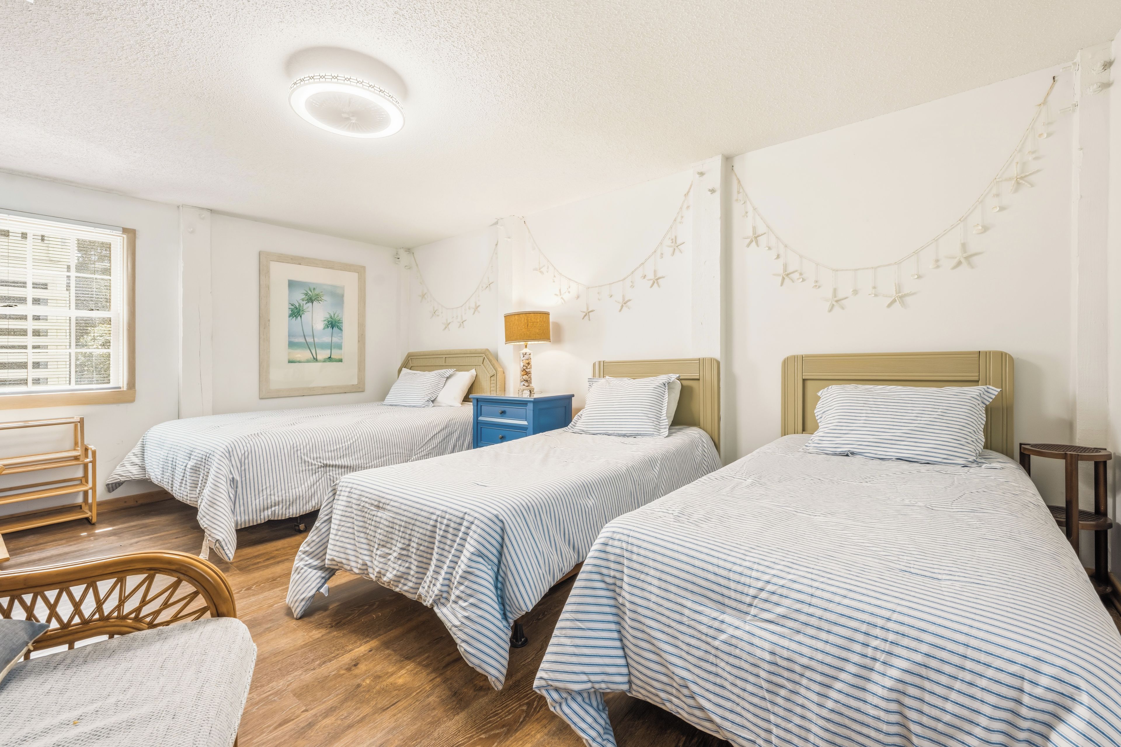 Une chambre avec trois lits simples, un plancher en bois, une fenêtre avec des stores, un tableau encadré au mur et une guirlande de décorations en forme d’étoiles.