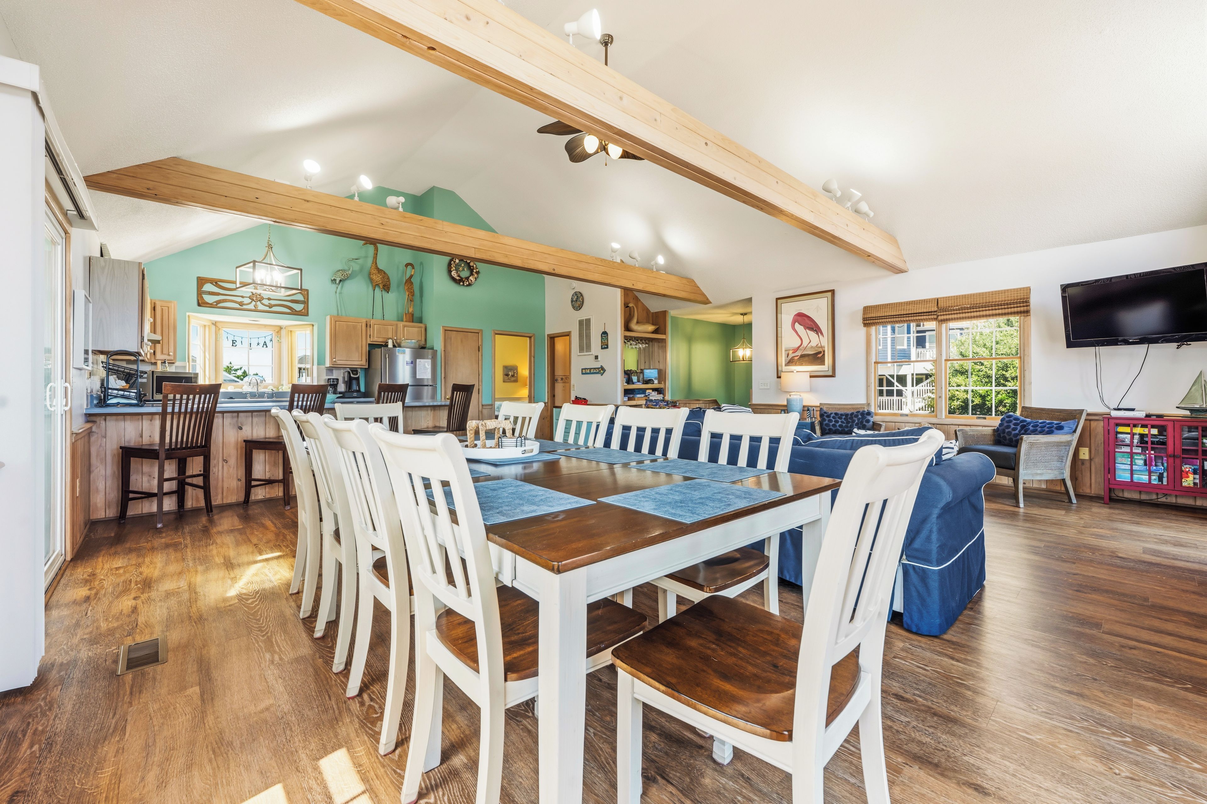 A spacious living area with a wooden dining table and chairs, a kitchen in the background, and a television mounted on the wall.