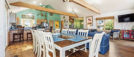 A spacious living area with a wooden dining table and chairs, a kitchen in the background, and a television mounted on the wall.