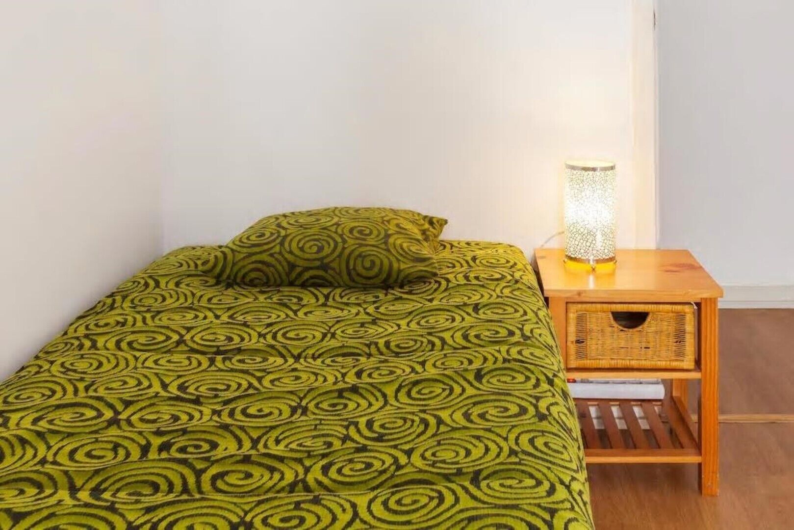A small bed with a green patterned bedspread and a matching pillow, a bedside table with a lamp, and a wicker basket under the table.